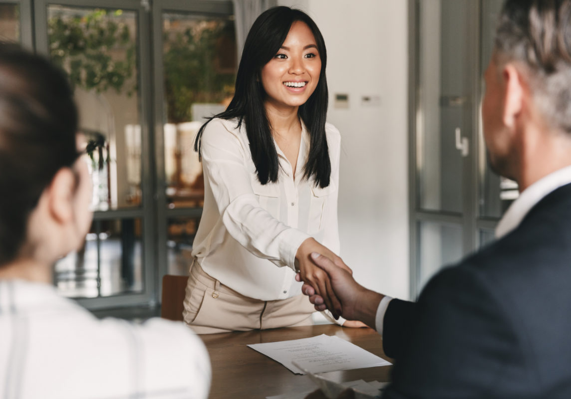 Business, career and placement concept - image from back of two employers sitting in office and shaking hand of young asian woman after successful negotiations or interview